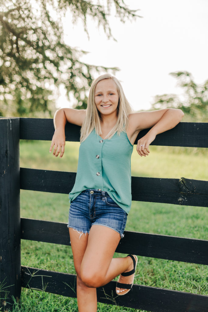 Augusta photography with senior photographer capturing girl in a green shirt and shorts leaning against a fence in the country of Georgia for her senior pictures 