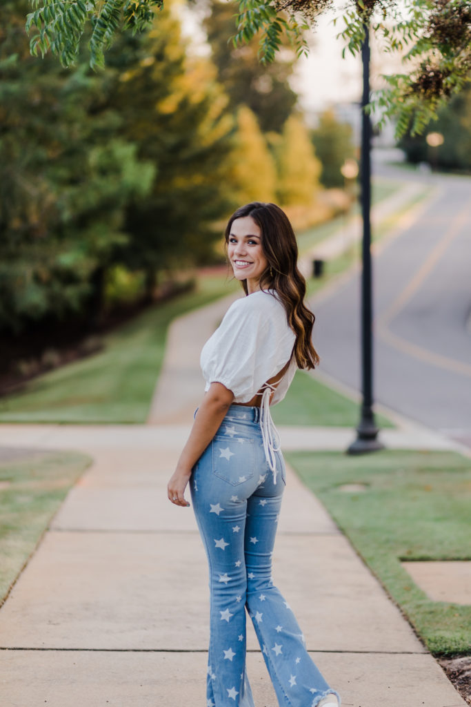 senior girl walking down the sidewalk in her neighborhood and turns around while the sun sets through the trees taken by augusta photographer