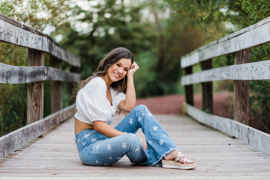 senior pictures with Augusta photographers with girl ina. white top and blue jeans sitting on a bridge for her fall senior pictures 