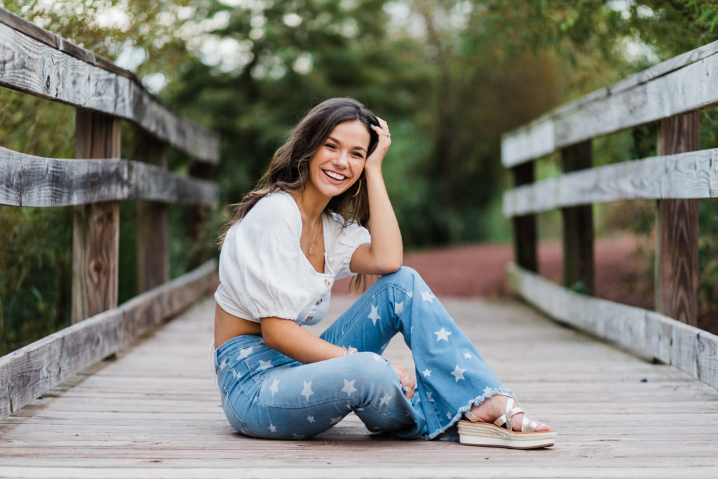 senior pictures on a lake with girl in a blue jeans with stars and a white top sitting on a bridge 