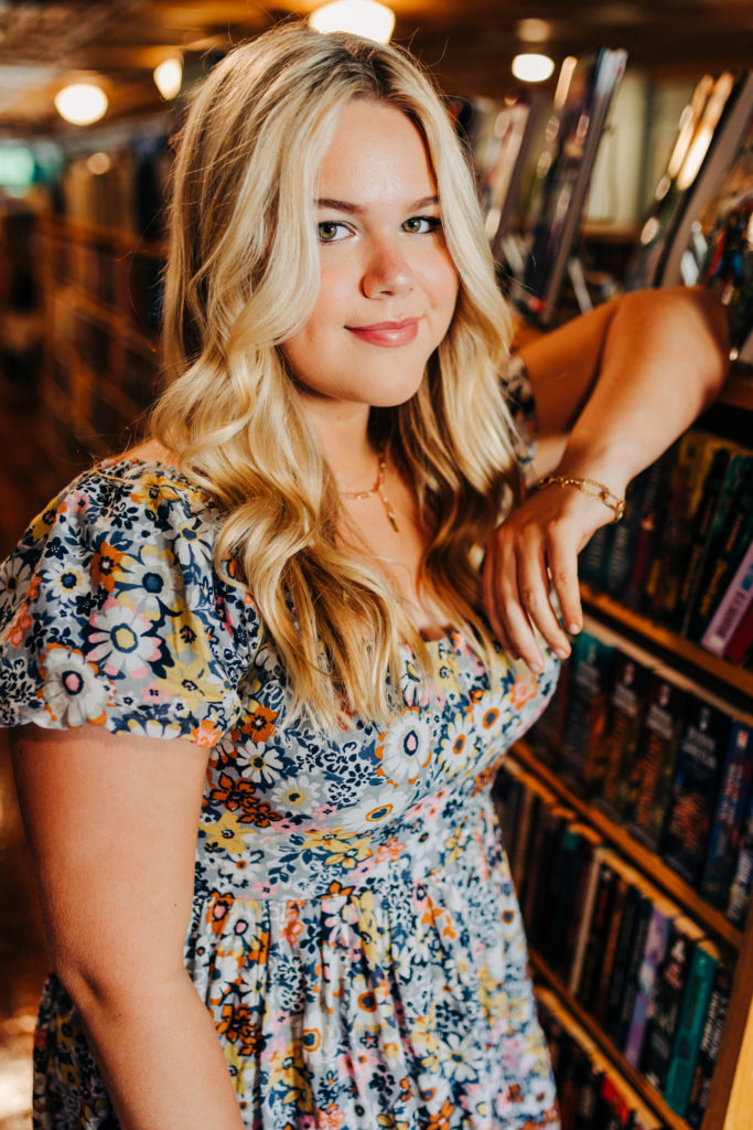 girl in a blue floral dress leaning against a book shelf for her senior photos