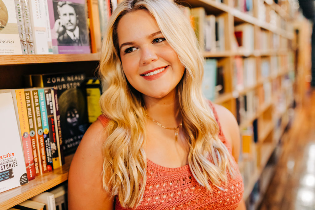 senior picture ideas with girl leaning against a bookshelf in a book store smiling 