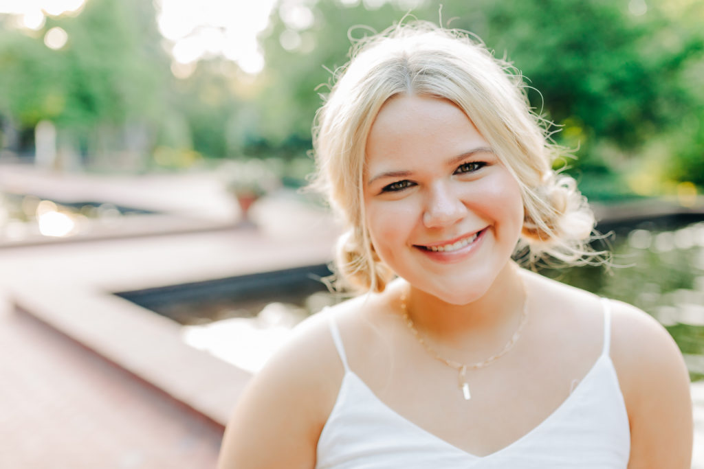 outdoor senior pictures with girl in a white dress smiling with a fountain behind her 
