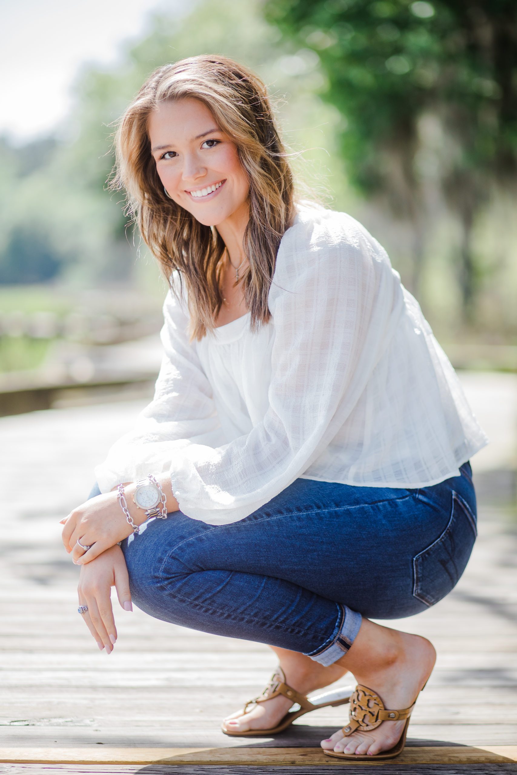 outdoor senior portraits with girl in blue jeans and a white blouse crouches down and smiles 