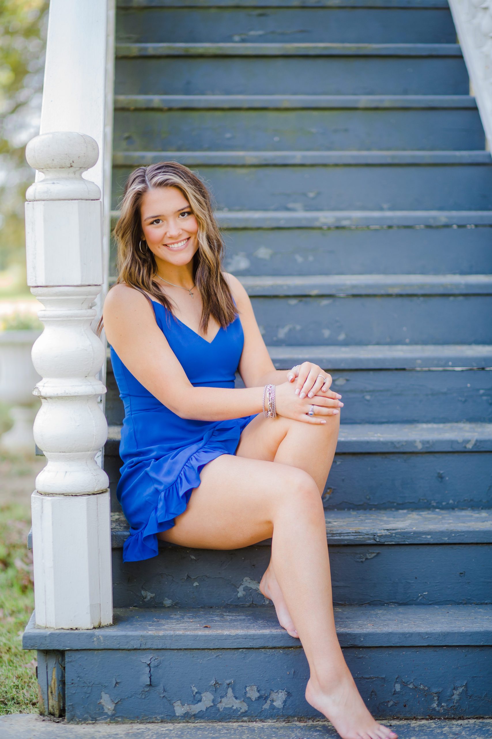senior girl in a little blue dress sits on a stair case outside of a home as she leans against the the railing 