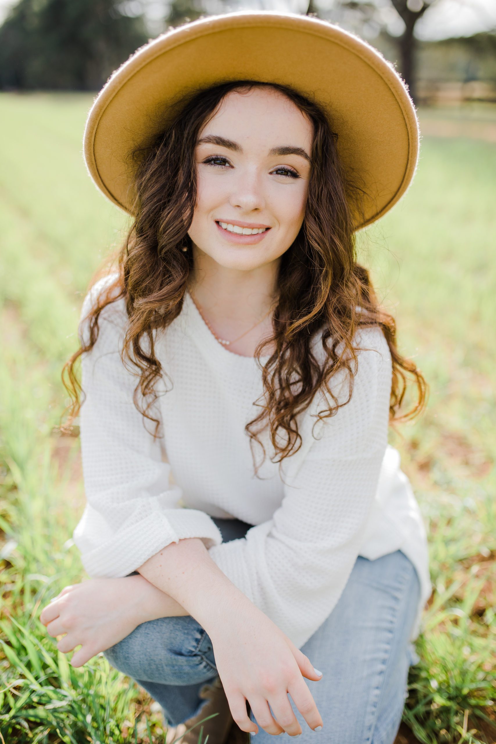 girl sitting on the ground ina green fiel in Georgia for her senior pictures as she wears a floral blouse and a wide brimmed hat 