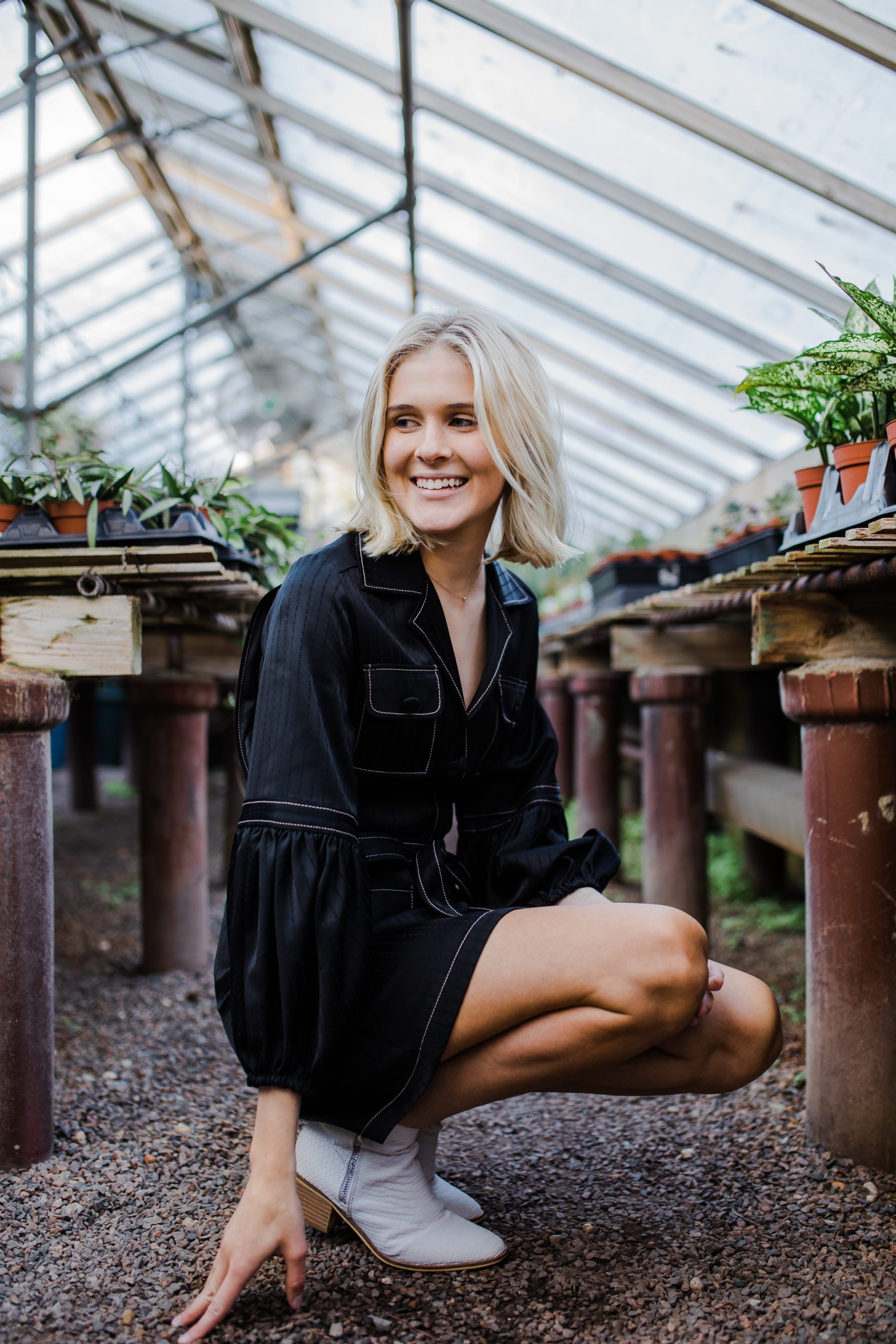 high school senior photographer captures young woman in a greenhouse in all black smiling as she crouches down 
