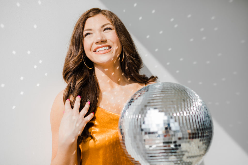 young woman posing for her senior photos while holding a disco ball and standing in the sunshine as the disco ball glitters in the light
